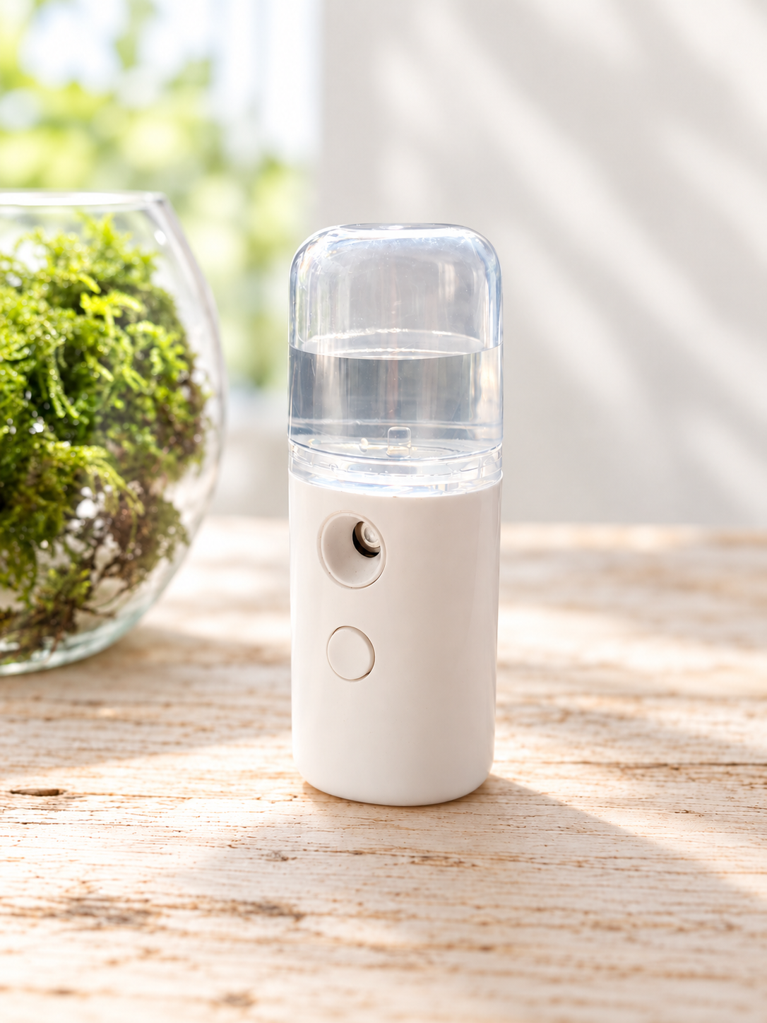 Compact white USB rechargeable plant mister beside a glass terrarium filled with fresh green moss